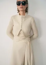 Jersey de punto acanalado en color beige, con cuello alto y cierre frontal de cremallera bidireccional. Presenta un diseño entallado de manga larga con una estructura que simula una prenda superpuesta.