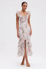 Vestido largo de corte midi con estampado floral en tonos suaves sobre base off-white. Presenta escote corazón con detalle de volantes en los hombros, cuerpo estilo corset y falda con volantes asimétricos y abertura frontal.