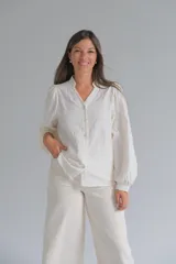 Blusa de color crudo con cuello mao y cierre frontal de botones. Presenta mangas largas abullonadas con puños y un delicado diseño bordado con relieve en toda la prenda.