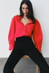 Camisa de popelina roja, de corte oversize, con cuello solapa, manga larga y bajo asimétrico redondeado. Cierre frontal oculto con botones.