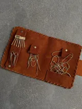 Set de accesorios compuesto por un sobre organizador de gamuza color marrón, un collar de cadena dorada, dos pares de caravanas colgantes y un prendedor, todos en metal dorado.