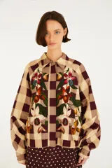 Chaqueta de cuello camisero, manga larga y cierre frontal oculto. Presenta un estampado de cuadros en tonos marrón y beige, complementado con un diseño bordado de flores y hojas en la parte delantera.