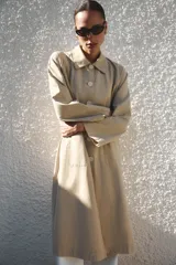 Trench largo de Zara Woman Collection, confeccionado en color beige. Cuenta con cuello de solapa, manga larga, bolsillos frontales de parche y cierre central con botones visibles.