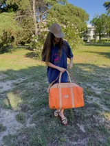 Bolso de viaje de lona color naranja con detalles en cuero tostado, asas cortas de mano y correa larga ajustable para el hombro.