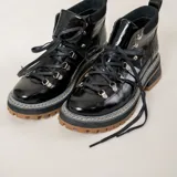 Borcegos de cuero charolado negro, de estilo montañero con cordones negros y ganchos metálicos. Presenta una suela gruesa track con plataforma y taco de goma marrón.
