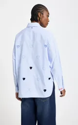 Camisa de algodón a rayas celestes y blancas, con cuello clásico, cierre frontal de botones, mangas largas y aberturas troqueladas en forma de corazón junto a bordados decorativos y texto en el hombro.