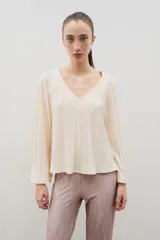 Blusa plisada de color crema con escote en V y mangas largas ligeramente acampanadas. Presenta una textura plisada en toda la prenda que aporta caída y movimiento.