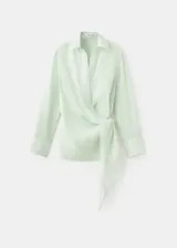 Camisa cruzada de ramio en color crudo, con cuello camisero, escote en V y mangas largas con puños anchos abotonados. Cuenta con un diseño envolvente con detalle de lazo lateral.