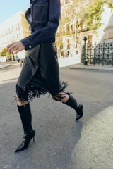 Falda midi negra con efecto piel, diseño cruzado en el delantero y flecos en el bajo. Cierre lateral con cremallera oculta.