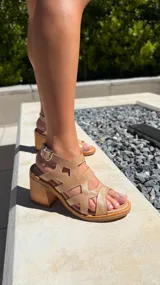 Sandalia dorada con tiras entrecruzadas con recortes en forma de rombo y taco ancho de madera. Posee cierre de hebilla en el tobillo.