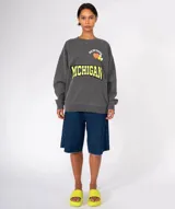 Buzo de lavado suave color mostaza con cuello redondo y manga larga. Presenta un estampado frontal con la palabra "MICHIGAN" en azul y amarillo, y apliques decorativos que incluyen un corazón rojo y dos pines circulares.