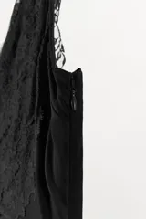 Vestido midi de viscosa color negro, sin mangas, con escote en pico adornado con encaje a tono. Presenta un detalle de nudo drapeado en la cintura y bajo asimétrico.