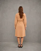 Vestido camisero color camel, de corte midi con mangas largas, cuello clásico, bolsillos frontales con solapa y cierre de botones. Incluye un cinturón de la misma tela para ajustar la cintura y presenta un ruedo redondeado con aberturas laterales.