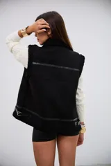 Chaleco negro de estilo aviador con interior y bordes de corderito sintético. Presenta cierre frontal metálico, detalles de tiras de cuero sintético y hebillas ajustables en el cuello y el ruedo.