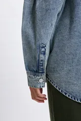 Sobrecamisa de jean en tono celeste con efecto lavado, corte oversize, cuello clásico, cierre frontal con botones metálicos y mangas largas con detalle de frunces en los hombros.