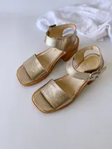 Sandalias de cuero color dorado, con tira ancha sobre los dedos, pulsera al tobillo con hebilla metálica y taco de madera.
