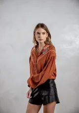 Camisa de color terracota con diseño de estilo western, presenta bordados blancos en la parte superior delantera y mangas largas abullonadas con puños ajustados. Cuenta con cierre frontal de botones y cuello camisero.