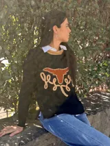 Sweater de punto fino color verde, con cuello redondo en contraste color beige. Presenta un diseño frontal con una figura de búfalo en cuero y detalles bordados a mano en color beige.