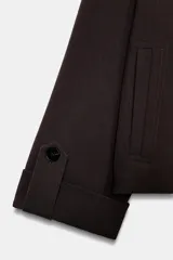 Trench corto de color marrón oscuro, con cuello de solapa, cierre frontal cruzado con botones y mangas tres cuartos con detalle de vuelta, trabilla y botón. Cuenta con bolsillos delanteros de vivo.