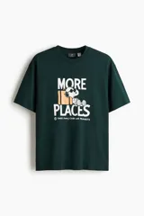 Remera verde oscuro con estampado frontal de Snoopy, equipaje y la inscripción "More Places - Take (more) care with Peanuts". Cuenta con cuello redondo y mangas cortas con hombros caídos.