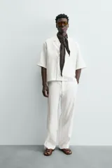 Camisa de lino color blanco, con cuello de solapas de muesca, manga corta y cierre frontal cruzado oculto. Presenta un calce relajado y aberturas laterales en el bajo.