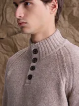 Sweater de punto en color beige, con cuello alto y cierre frontal parcial de botones. Presenta mangas largas con puños acanalados y diseño de corte clásico.