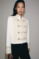Chaqueta de estilo militar de ZARA Woman Collection, confeccionada en mezcla de algodón en color blanco y beige. Presenta cuello subido, manga larga con puños anchos en contraste y cierre frontal con alamares y botones metálicos en relieve. Tiene bajo asimétrico con abertura en la espalda.