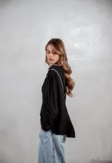 Camisa negra de manga larga con bordados blancos en la parte superior delantera y hombros. Presenta cuello mao, cierre frontal con botones y mangas con puños ajustados.