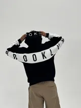 Canguro con capucha de corte boxy, color negro con una franja horizontal blanca que recorre la espalda y las mangas, presentando la palabra 'BROOKLYN' estampada en negro sobre la franja. La capucha también incluye detalles estampados.