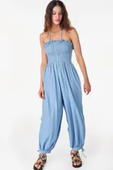 Mono largo de denim color celeste, sin mangas, con escote nido de abeja elástico y volado superior, tirantes finos para atar al cuello, perneras amplias de corte abombado con puños ajustables mediante lazos y bolsillos laterales.
