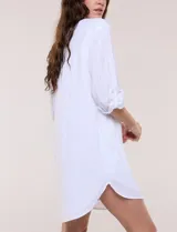 Vestido camisero corto de color blanco con sutiles rayas verticales, de la marca Dash. Presenta cuello con solapa, escote en V con botones parciales en el frente, bolsillo superior en el pecho y bolsillos laterales de gran tamaño. Cuenta con mangas largas de largo regulable mediante presilla y botón, calce holgado y ruedo trasero más largo.