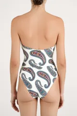 Malla enteriza de color blanco con estampado paisley en tonos azules, marrones y rojos. Presenta diseño bandeau con tirantes finos ajustables, escote con aberturas circulares unidas por argollas metálicas doradas.
