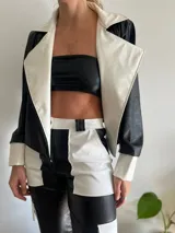 Chaqueta de cuero sintético negra con detalles en blanco, de corte recto y solapas amplias. Incluye un top bandeau negro y un pantalón bicolor de cuero sintético negro y blanco.