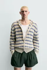 Cárdigan de punto con diseño de trenzas y rayas horizontales en tonos beige y azul. Presenta corte relajado, capucha, cierre frontal con cremallera y puños y ruedo en tejido rib.