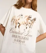 Remera oversized de algodón color crudo, con cuello redondo y mangas cortas. Estampa trasera con texto "Amor Peligroso" y listado de ciudades con fechas, en tipografía estilo western.
