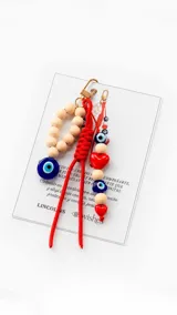 Cartera tipo hobo de cuero sintético color suela, con interior espacioso y costuras decorativas frontales. Incluye asa de hombro fija, monedero rectangular colgante a juego y un llavero amuleto con cuentas de madera, ojos turcos y detalles en rojo.