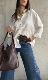 Camisa oversize blanca con botones frontales, cuello camisero y manga larga. Presenta grandes bordados en hilo beige con diseño de hojas o abanicos en la parte inferior frontal.