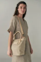 Cartera tipo bolso de mano confeccionada en lana fina sin teñir, con detalles en cuero marrón oscuro en las asas y correa ajustable. Incluye cierre superior y etiqueta de cuero con logo.