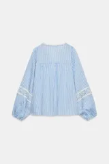 Blusa de rayas verticales blancas y celestes, con cuello redondo y escote en pico. Presenta detalles de bordados perforados y puntillas en el pecho y las mangas. Tiene mangas largas abullonadas con puños elásticos y cierre frontal con botones forrados.