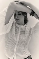 Camisa blanca de algodón con cuello alto y volados, mangas largas abullonadas con puños elásticos. Presenta detalles de encaje y frunces en el frente y la espalda.