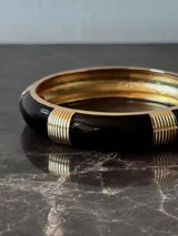 Pulsera rígida tipo brazalete, color negro con detalles dorados.
