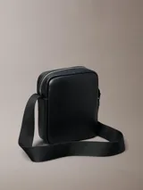 Cartera reporter estructurada Calvin Klein de poliuretano con textura granulada, color negro, cierre superior de cremallera y bolsillo frontal con cierre. Cuenta con correa de hombro ajustable y logo metálico de la marca en la parte delantera.