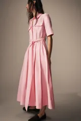 Vestido midi de estilo camisero color rosado, con cuello solapa, manga corta y cierre frontal con botones. Presenta bolsillos de plastrón con solapa en el pecho, bolsillos laterales ocultos y un cinturón del mismo tejido para ajustar en la cintura.