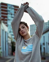 Sweater de hilo de algodón en color beige con diseño tejido de la bandera de Uruguay en el centro. Presenta cuello redondo, mangas largas y un corte holgado.