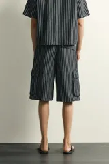 Bermuda cargo relaxed fit confeccionada en denim de mezcla de algodón y lino, con diseño de rayas verticales grises y negras. Cuenta con bolsillos laterales tipo cargo con solapa y cierre frontal con cremallera y botón.