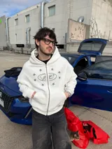 Campera canguro blanca con cierre frontal, capucha y bolsillos tipo kankaro. Presenta un estampado frontal con un óvalo con texto cursiva rodeado de estrellas negras.