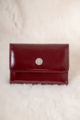 Billetera de cuero charolado color bordó con solapa frontal y aplique metálico circular con logo grabado.