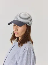 Gorro tipo pañuelo con visera rígida azul marino, confeccionado en lino con estampado de rayas grises y blancas. Presenta un diseño envolvente con caída trasera y detalle de logo bordado en el lateral.