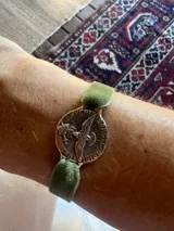 Pulsera con cinta de terciopelo verde y dije redondo plateado con grabado de un águila y texto en latín.