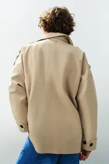 Trench corto oversize color beige con cuello subido de efecto doble, cierre cruzado con botones oscuros y bolsillos de vivo delanteros.